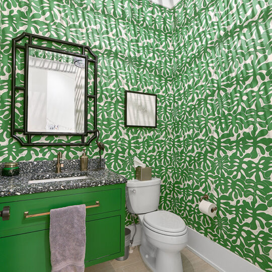 A bathroom features bright green and white patterned wallpaper with leafy designs. It has a green vanity with a dark countertop, a rectangular mirror, a toilet, and a modern black hanging light fixture casting shadows on the ceiling.