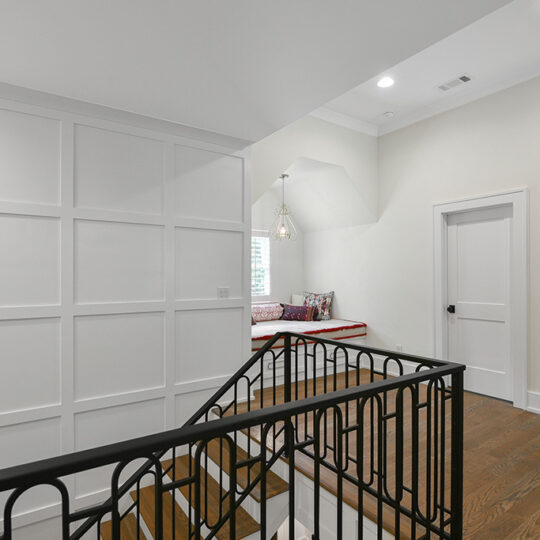 A staircase with a black metal railing leads to an upper hallway with wooden floors. A white paneled wall is on the left. A cozy window seat with colorful cushions is in the corner, beneath a small hanging lamp. A door and framed artwork are on the right.