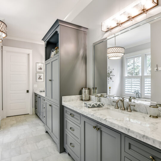 Modern bathroom with gray cabinetry, marble countertops, and a large mirror. Features two stylish light fixtures, decorative wall art, and a patterned rug on the marble floor. A window with shutters lets in natural light.