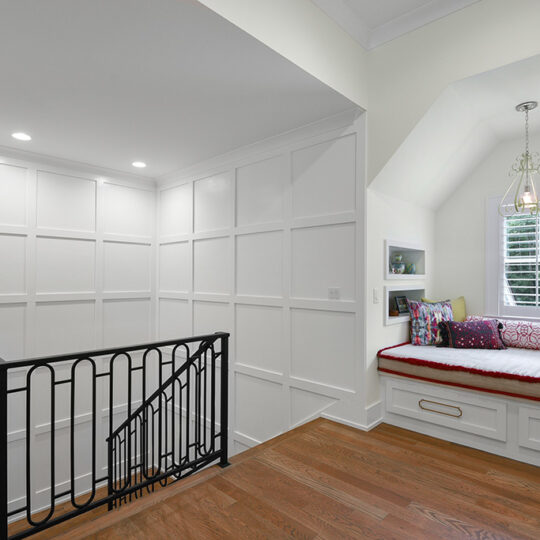 A bright hallway features a white paneled wall, a black metal railing, and wooden flooring. To the right, theres a cozy window seat with colorful cushions and built-in drawers beneath a chandelier-style light fixture.