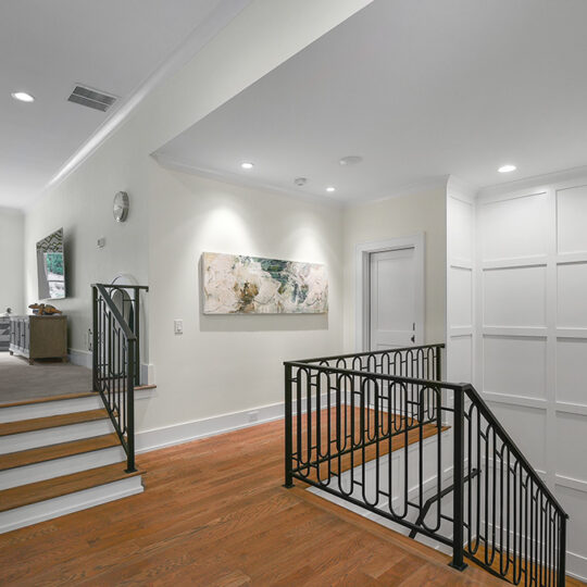 A modern interior featuring a staircase with dark metal railing, leading up to a spacious, well-lit room. Hardwood floors complement the light-colored walls, adorned with a piece of abstract art. Large windows and recessed lighting illuminate the space.