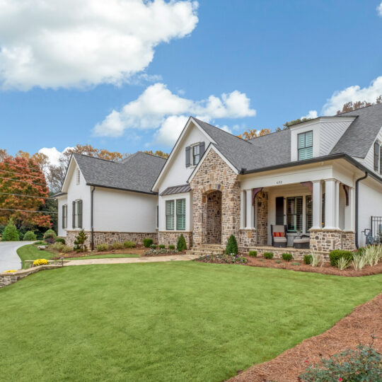 A charming suburban house with beige brick and white siding is surrounded by a well-maintained green lawn. It features a front porch with red chairs and is set against a backdrop of trees under a partly cloudy sky.