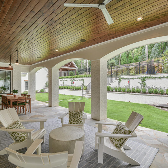 A covered patio with a wooden ceiling and a ceiling fan. Four white Adirondack chairs surround a round table on a grey rug. A dining table is in the background, and a green egg grill is to the right. Lush greenery and trees are visible beyond the patio.