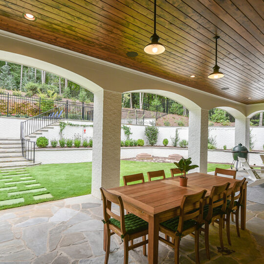 A spacious covered patio with a wooden ceiling, hanging lights, and a large table with chairs. A stone fireplace and seating area are at one end. The patio overlooks a landscaped garden with a stone path, steps, and lush greenery.