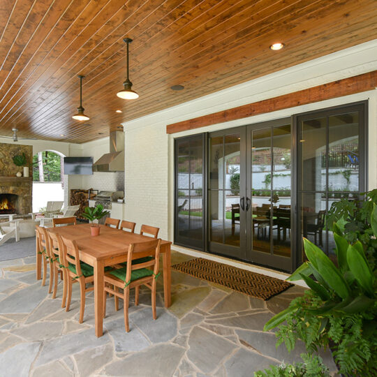 Outdoor patio with a wooden ceiling and hanging lights, featuring a wooden dining table with chairs on a stone floor. Theres a fireplace with stone surround, plants, a rug inside sliding glass doors, and a grill on the left.