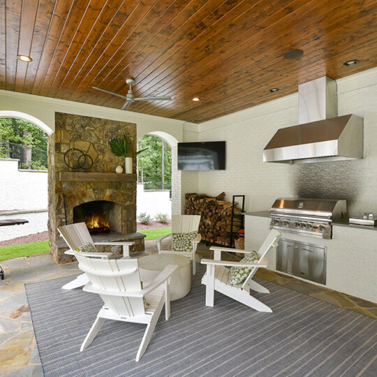 Outdoor patio with a wooden ceiling, a fireplace, and a grill. Three white chairs are around a round table on a striped rug. Theres a green egg-style barbecue, a stack of firewood, a wall-mounted TV, and a ceiling fan. Stone flooring completes the space.