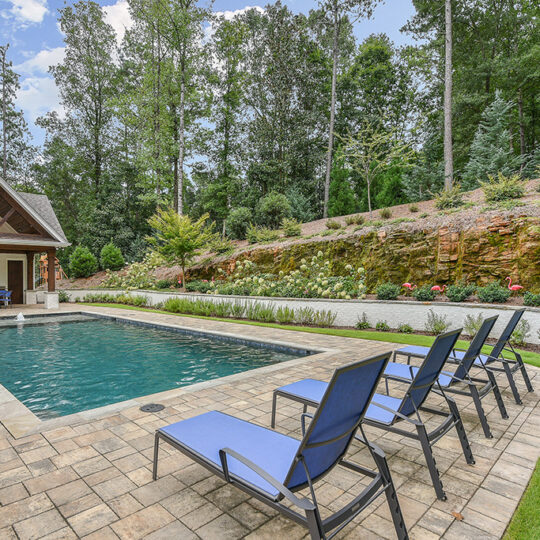 A serene backyard scene with a rectangular swimming pool surrounded by stone tiles. Four blue lounge chairs are lined up on one side. A small pavilion with a peaked roof is in the background, set amidst lush greenery and trees.