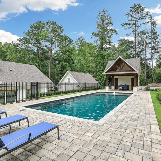 A rectangular swimming pool is surrounded by a stone patio with a line of blue lounge chairs. A covered seating area is at one end, and the pool is bordered by a black fence and greenery. Trees and a house are in the background.