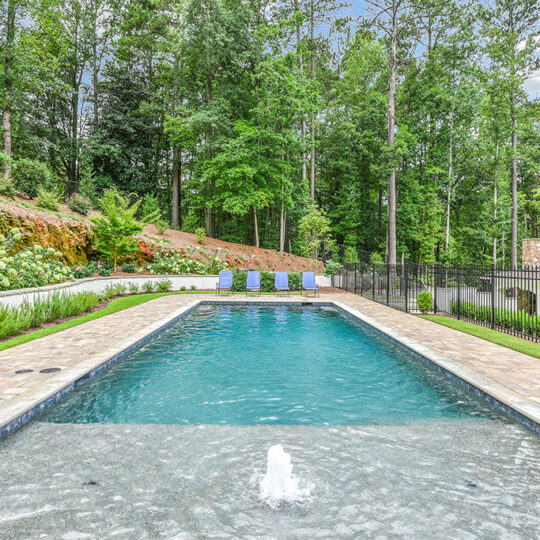 Outdoor rectangular swimming pool surrounded by stone tiling. A fountain sprays water at one end, and four blue lounge chairs sit at the other. Tall trees and greenery border the pool area, with a metal fence and a house visible in the background.