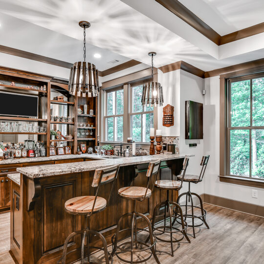 Rustic home bar with wooden cabinets, a granite countertop, and four metal stools. Shelves are stocked with bottles and decor. Pendant lights hang from the ceiling. Large windows offer a view of lush greenery outside.