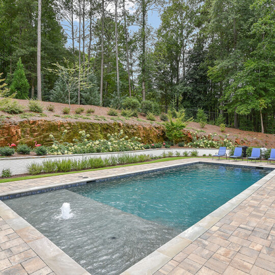 A rectangular pool with a fountain feature is surrounded by a paved patio. Four blue lounge chairs are placed to the right. In the background, theres landscaping with greenery, a stone wall, and trees.