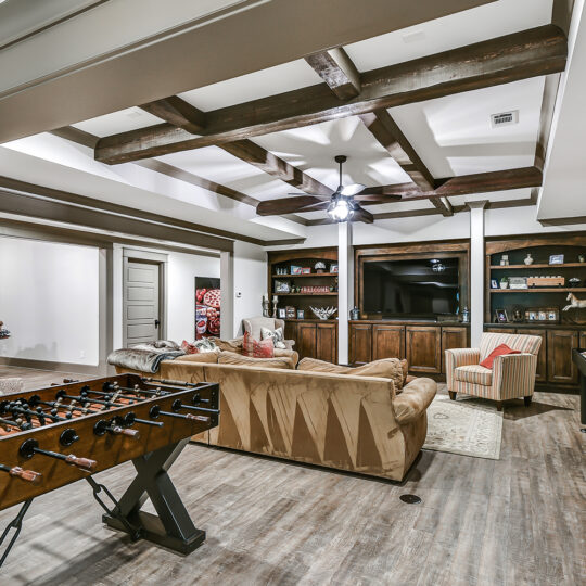 A spacious, modern entertainment room with a foosball table and shuffleboard. A sectional sofa and armchair face a large wooden entertainment center with a TV and shelves. Exposed wooden beams on the ceiling complete the cozy ambiance.