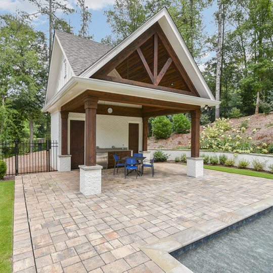 A backyard scene with a small covered pavilion on a patio adjacent to a rectangular pool with a fountain. Blue chairs and a table sit under the wooden pavilion. Trees and greenery surround the area, with a metal fence on the left.