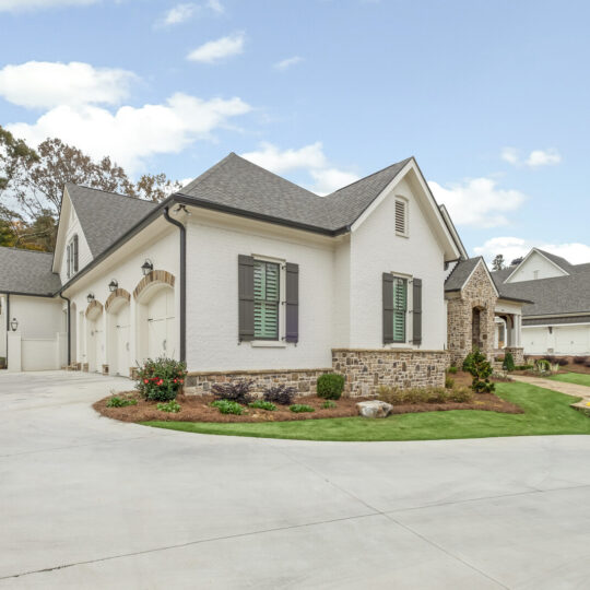 A large, elegant house with white brick, grey shutters, and a steep, dark grey roof. It features multiple garage doors and a landscaped front yard with shrubs and a stone retaining wall. The sky is partly cloudy.