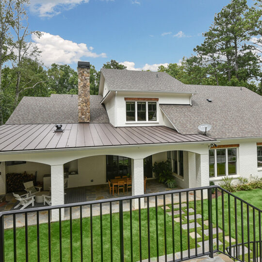 Backyard view of a modern house with white walls and a dark gray roof, featuring a covered patio with seating, surrounded by a well-manicured lawn. Tall trees are in the background under a partly cloudy sky.