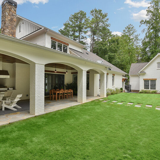 Spacious backyard with white brick patio featuring an outdoor dining area and lounge chairs. Green lawn with stepping stones leads to a separate white building. Surrounded by lush trees under a partly cloudy sky.