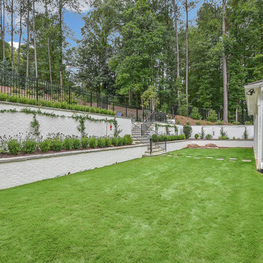 A well-maintained backyard with a lush green lawn, bordered by white retaining walls with plants. Theres a metal staircase leading to an upper garden area, surrounded by tall trees. A white house is visible on the right.