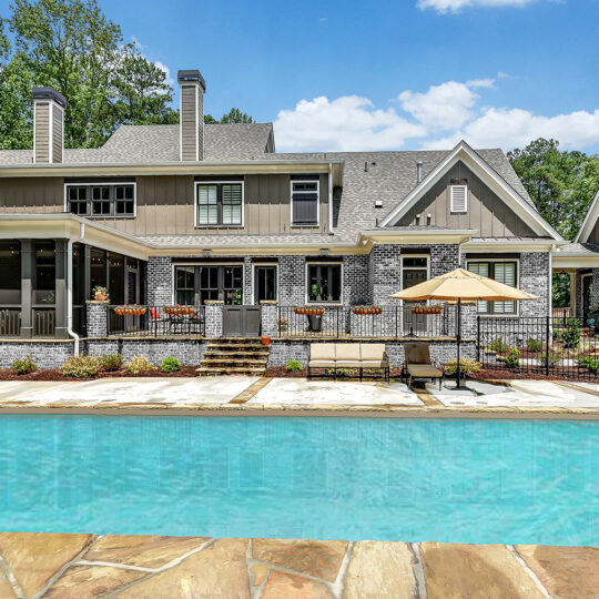 A large, elegant house with a mix of stone and wood siding sits behind a clear blue swimming pool. Patio furniture with an umbrella is arranged poolside. The home features tall windows and a covered porch, surrounded by lush greenery and trees.