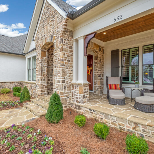 A cozy, stone-clad house features a front porch with two cushioned chairs and colorful striped pillows. The entrance is detailed with wooden beams. The yard is neatly landscaped with small shrubs and flowers under a bright, partly cloudy sky.