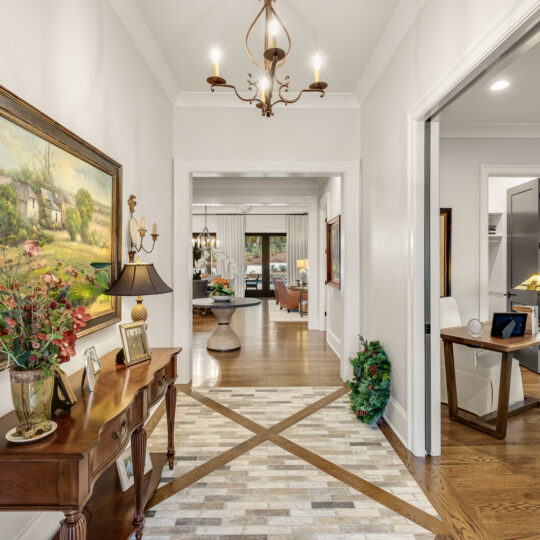 A well-lit hallway with a wooden console table on the left, adorned with a lamp, flowers, and paintings. The floor is tiled in a geometric pattern. The hallway leads to a bright living area with a dining table visible in the background.