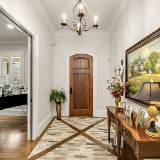 A hallway with a wooden door, a painting, a table with family photos, and a lamp. The floor is tiled with a geometric pattern. To the left is an open room with a black grand piano and large windows with shutters. A chandelier hangs from the ceiling.