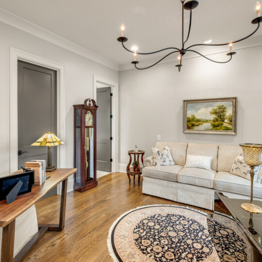 A cozy living room with a white sofa, a glass coffee table, and a patterned rug. A tall grandfather clock stands near a door. A chandelier hangs from the ceiling. Framed art and family photos decorate the walls and table. A green plant is in the corner.