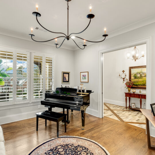 A music room with a black grand piano, surrounded by framed artwork. A round rug is on the wooden floor, and a couch with a patterned pillow is nearby. A large plant is in a corner, and a modern chandelier hangs from the ceiling.