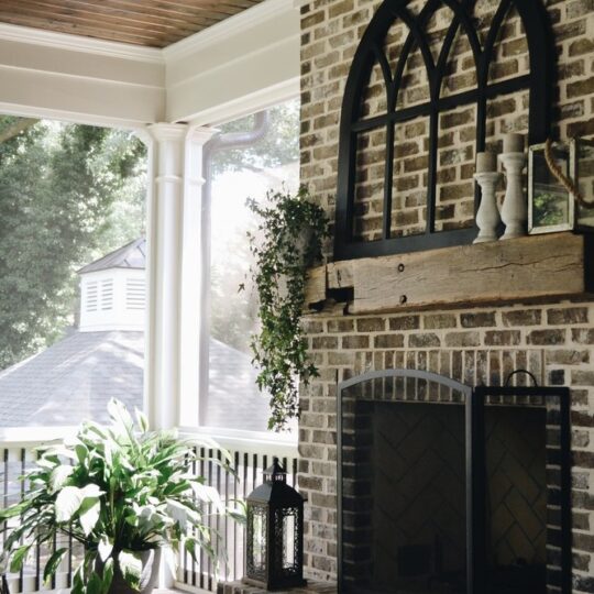 A cozy porch with a brick fireplace adorned with decorative items and plants. Sunlight filters through large windows, casting shadows on the floor. A roof is visible through the window, surrounded by greenery. Nearby chairs and potted plants create a serene ambiance.