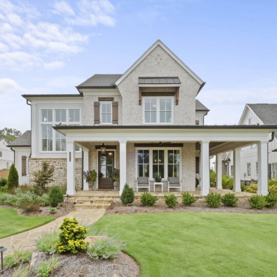 A modern two-story house with a gabled roof, large windows, and a covered porch. The exterior is a combination of light brick and siding. Lush green lawn and landscaped shrubs are in the front yard, under a partly cloudy blue sky.