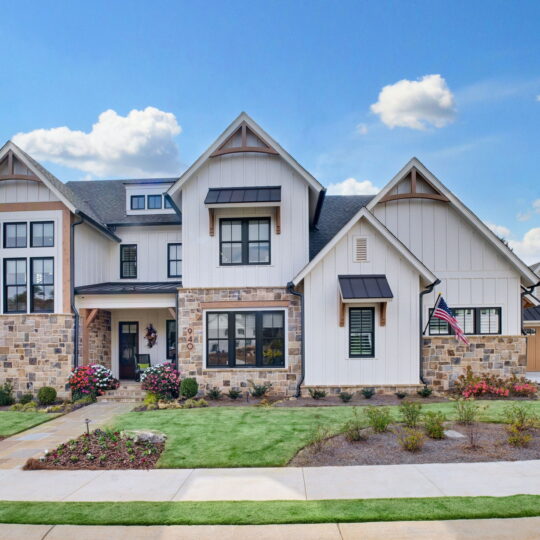 A modern suburban house with a mix of stone and white siding, featuring multiple peaked rooflines. The front yard is landscaped with flowers and a lawn. An American flag is displayed near the entrance. The sky is partly cloudy.