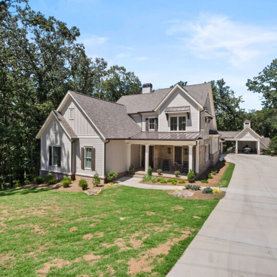 A modern two-story house with a large, sloping roof, surrounded by trees. The house has beige siding and a covered porch. A long concrete driveway leads to a carport. The front yard is partially grassed with landscaped bushes.