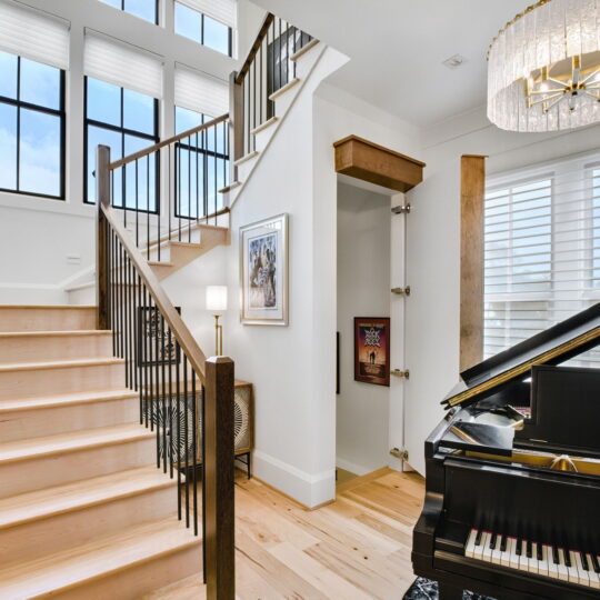 Spacious modern interior featuring a light wooden staircase with black railings, large windows with blinds, a chandelier, and a black grand piano on a wooden floor. A framed picture and decorative elements adorn the white walls.