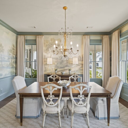 Elegant dining room with a wooden table and six chairs, featuring a chandelier above. Large windows with cream curtains allow natural light. Walls are adorned with pastoral landscape murals, complementing light blue paneling and a patterned rug.