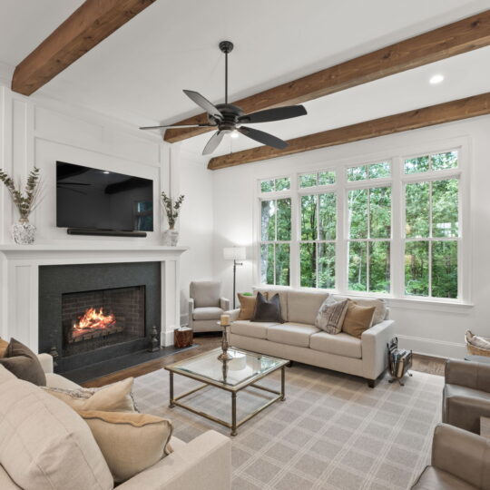 A cozy living room with a fireplace and wall-mounted TV. It features a gray sofa set, armchairs, and a glass coffee table on a patterned rug. Wooden beams accent the ceiling, and large windows offer views of green foliage outside.