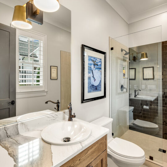 A modern bathroom features a marble countertop with a white vessel sink and a wooden cabinet. On the wall, theres a framed picture and two gold light fixtures. The glass-enclosed shower has beige tiles, and there is a toilet nearby.