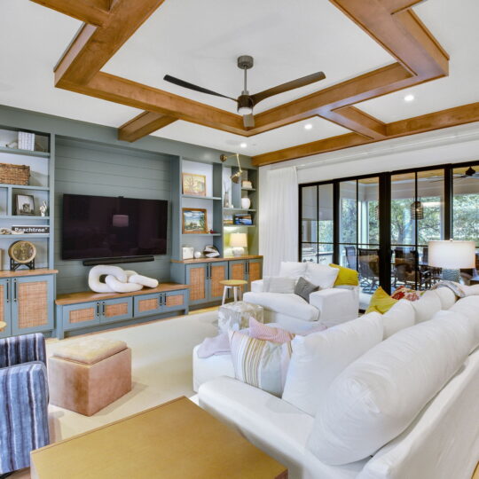 Spacious living room with a modern design featuring a wooden ceiling accent, ceiling fan, large TV on a built-in shelf unit, and white sofas. A striped armchair and pouf complete the seating. Floor-to-ceiling windows showcase a view of a porch and trees.