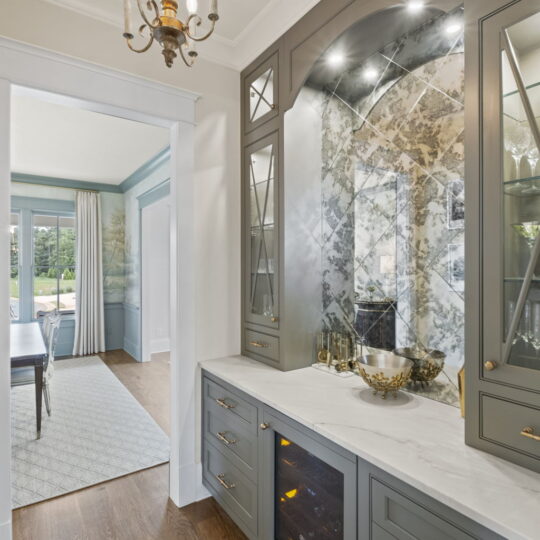 Elegant hallway with a gray cabinet, white countertop, and mirrored backsplash. A chandelier hangs from the ceiling. The adjacent room has a dining table, chairs, and large windows with curtains, revealing a view of greenery outside.