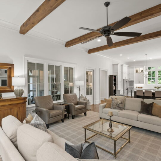 A spacious, modern living room with white walls, wooden ceiling beams, and hardwood floors. It features a beige sofa, armchairs, a coffee table, a wooden sideboard with a mirror, and a ceiling fan. The kitchen is visible in the background.