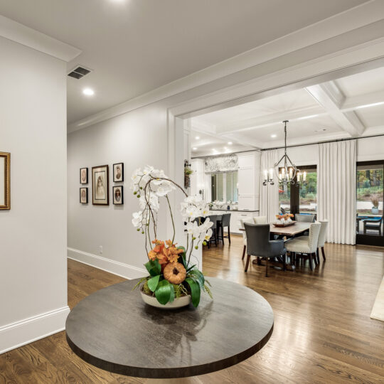 Spacious open-concept living and dining area with wooden floors. A large round table holds an orchid arrangement. The dining area has a table, chairs, and a chandelier. Glass doors lead to an outdoor space. Framed art decorates the walls.