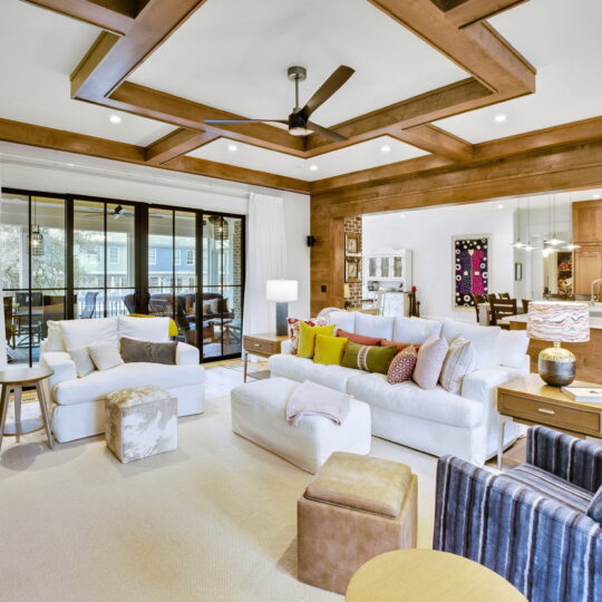 Modern living room with white sofas, colorful cushions, and wooden beams on the ceiling. A glass door opens to an outdoor area, and the space connects to a kitchen with a bar counter. Bright and inviting with a blend of textures and decor.