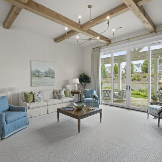 A spacious living room with a beige couch, two blue armchairs, and a wooden coffee table. Large windows reveal a garden view. The ceiling features wooden beams, and elegant curtains frame the glass doors. A landscape painting decorates the wall.