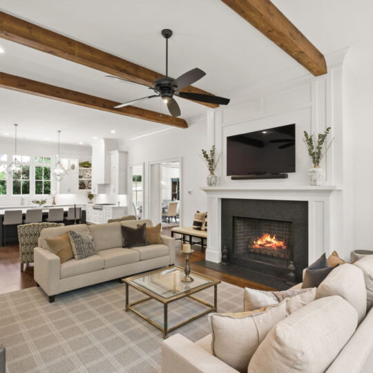 Spacious living room with beige sofas and a coffee table centered on a checkered rug. A fireplace with a mounted TV is on one wall. The room is open to a modern kitchen with white cabinets and a dining area. Exposed wooden beams line the ceiling.