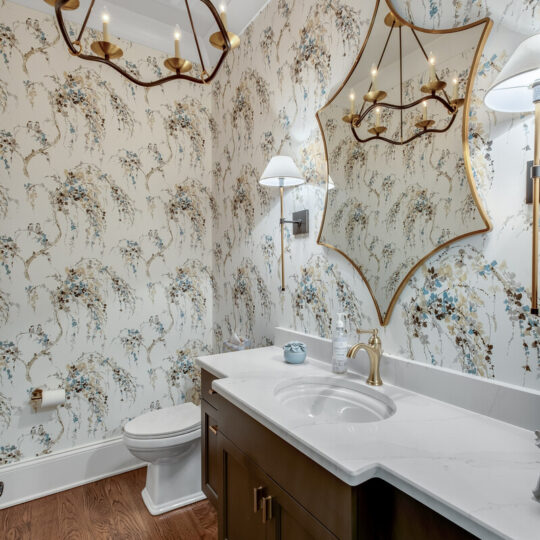 Elegant bathroom with patterned floral wallpaper, a star-shaped mirror, and a modern light fixture with candle-shaped bulbs. The wooden vanity features a white marble countertop, gold faucet, and there are wall sconces flanking the mirror.