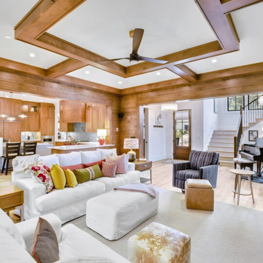 Spacious open-concept living room with white sofas, colorful cushions, and wooden accents. A ceiling fan and recessed lighting illuminate the space. In the background, theres a dining area, kitchen, and a black grand piano near a staircase.