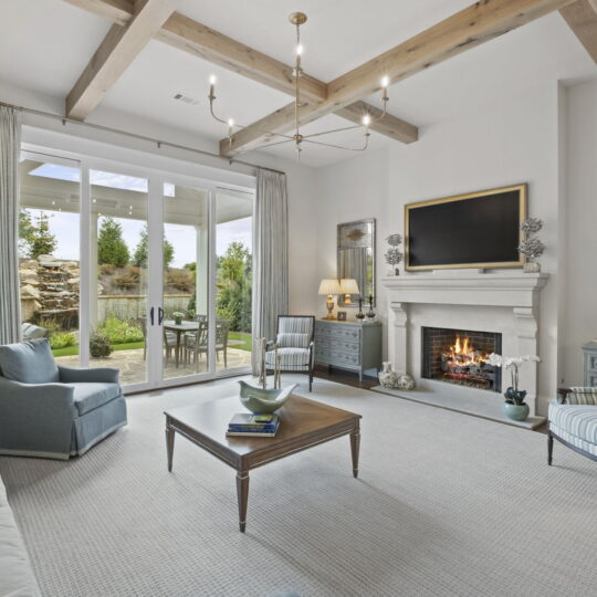 A cozy living room with a modern design features a fireplace, cushioned seating, and a TV above the mantel. Large glass doors open to a garden view, while a wooden chandelier hangs from the ceiling with exposed beams. Decor includes lamps and mirrors.