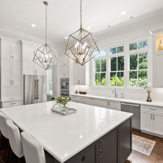 Modern kitchen with a large white marble island, three cushioned stools, and geometric pendant lights. White cabinets and stainless steel appliances line the walls. Large windows provide ample natural light, and hardwood floors add warmth.