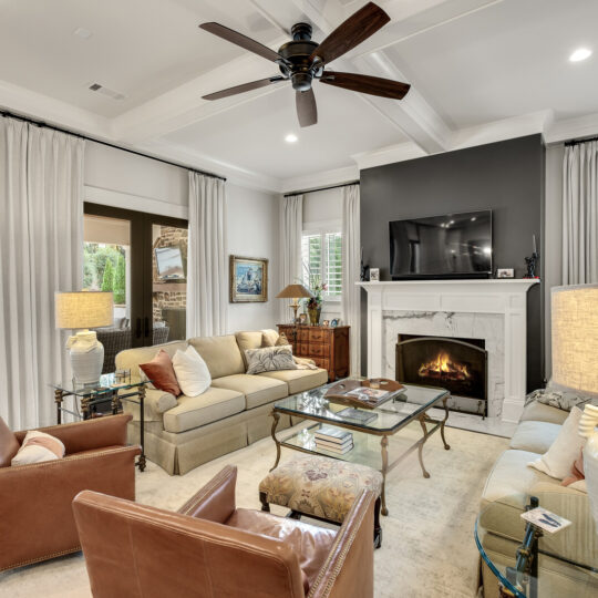 A cozy living room with beige sofas and brown leather chairs arranged around a glass coffee table. A fireplace with a TV above it is centered on a wall. Large windows with curtains and a ceiling fan enhance the airy atmosphere. Sliding doors lead to a patio.