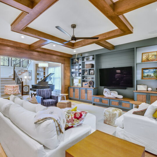 Modern living room with white sofas, a striped armchair, and a wooden coffee table. Large TV on a built-in shelf with decor items. Wooden ceiling beams, a ceiling fan, and a grand piano by the staircase. Bright, ambient lighting enhances the cozy space.