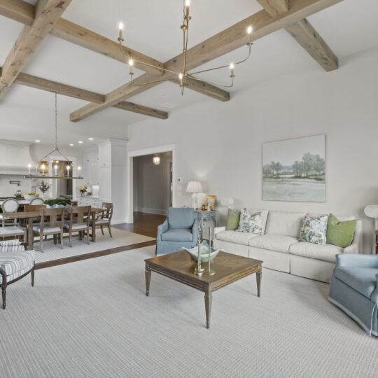 A bright and spacious living room features wooden ceiling beams, a chandelier, and white walls. It includes a sofa, two blue chairs, a wooden coffee table, and a dining table in the background. A painting and lamps add decor. Hardwood floors are partially covered by a rug.