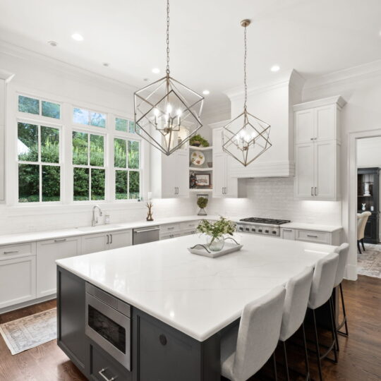 A modern kitchen featuring a large island with a white countertop and four chairs, pendant lights, white cabinets, and stainless steel appliances. A window above the sink overlooks greenery, and dark hardwood floors complete the design.